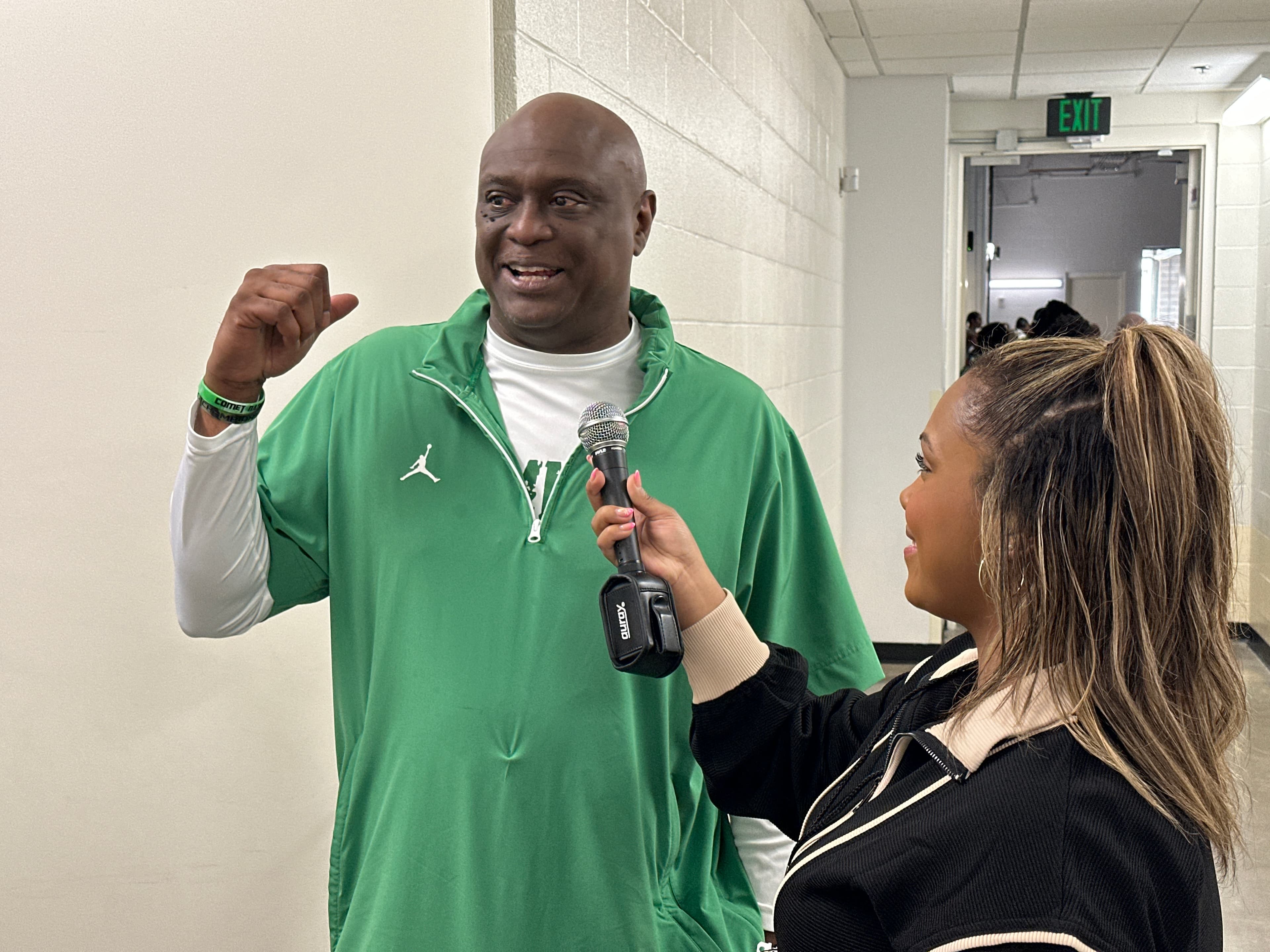 A coach in a green pullover speaks into a microphone during a hallway interview.