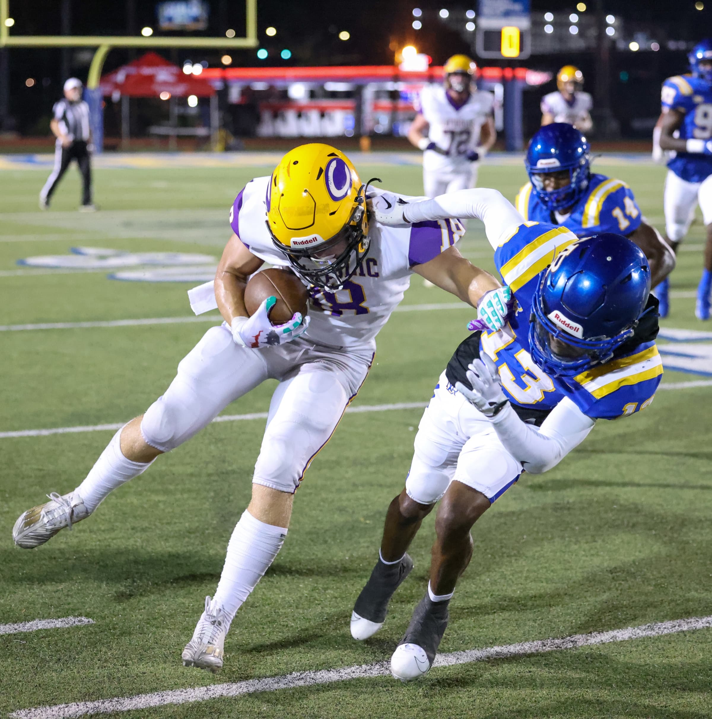 A high school football ball carrier fights through a tackle under stadium lights.