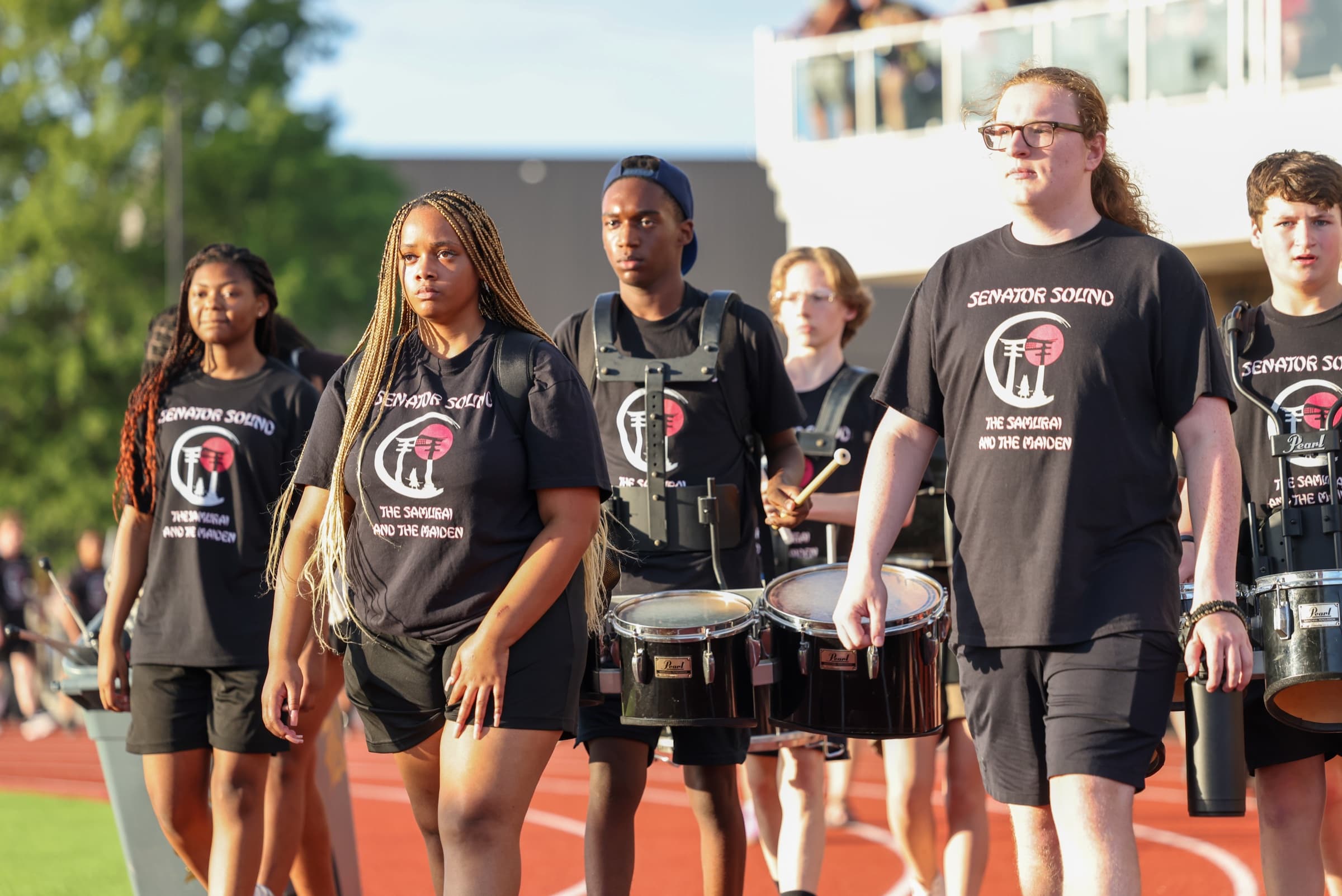 Student percussionists stand with marching drums beside a high school football field.